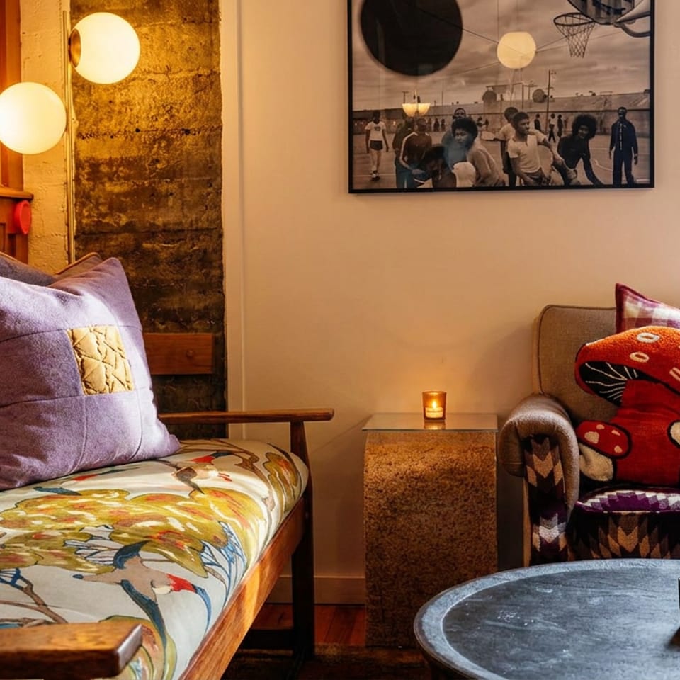 Cozy restaurant lounge with colorful upholstered seating, mushroom-shaped cushion, candles, artwork, and large windows.
