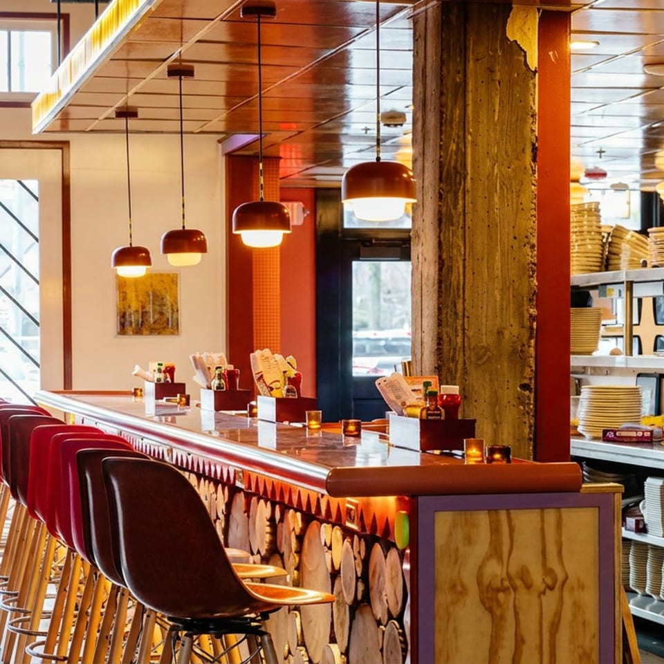 Warmly lit restaurant dining counter with red bar stools, pendant lights, wood accents, and an open kitchen stacked with plates.