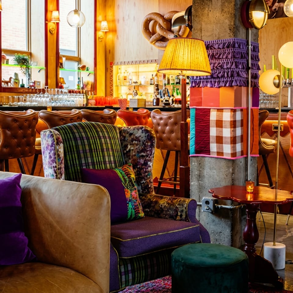 Colorful restaurant bar lounge with tufted leather stools, plush patterned seating, vibrant pillows, warm lighting, and liquor shelves.